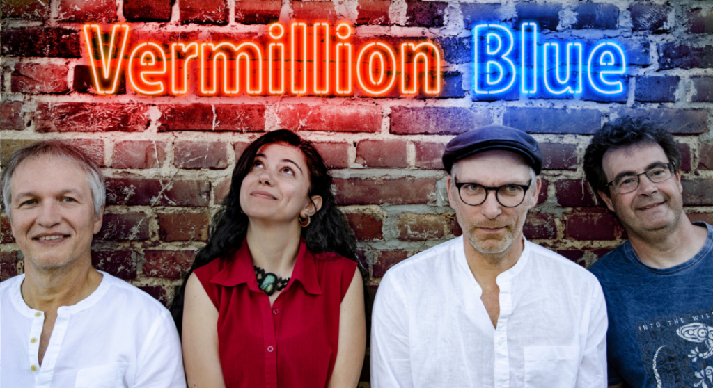 Die vier Bandmitglieder stehen vor einer Backsteinwand, über ihnen ein Neonschriftzug "Vermillion Blue"